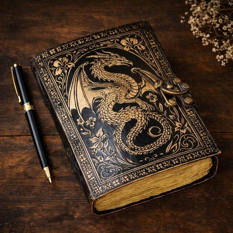 May include: A black leather journal with a gold embossed dragon design and floral accents. The journal has a clasp closure and gold-edged pages. A black and gold pen rests beside the journal on a wooden surface.