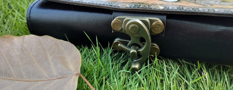 May include: A black leather clutch with a brass clasp and decorative stitching. The clutch is lying on a bed of green grass.