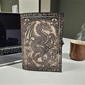 May include: A leather-bound journal with a detailed dragon illustration. The cover features a carved dragon design with floral accents. The journal is closed with a metal clasp. A brown mug with a snake skin pattern sits nearby.