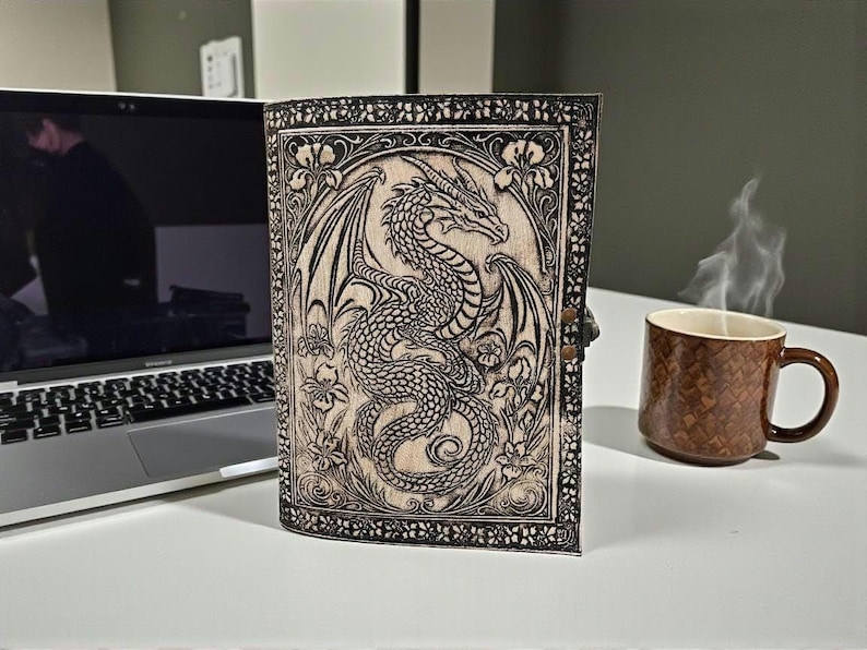 May include: A leather-bound journal with a detailed dragon illustration. The cover features a carved dragon design with floral accents. The journal is closed with a metal clasp. A brown mug with a snake skin pattern sits nearby.