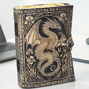 May include: A black leather journal with a dragon embossed on the cover. The dragon is tan with intricate details, surrounded by floral designs. The journal has a metal clasp and a black spine.