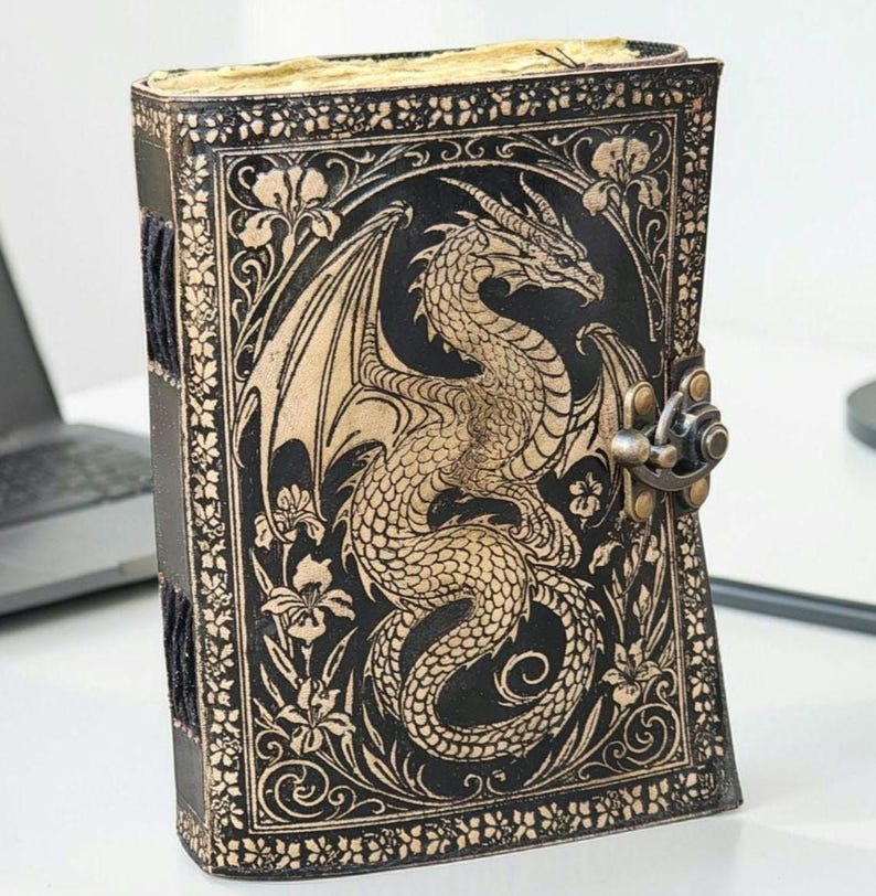 May include: A black leather journal with a dragon embossed on the cover. The dragon is tan with intricate details, surrounded by floral designs. The journal has a metal clasp and a black spine.