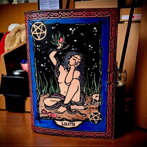 May include: A leather-bound journal featuring a detailed illustration of Lilith. The artwork depicts a woman with long hair, a snake, and a starry night background. The journal has a blue border and red and brown accents, with the name "LILITH" at the bottom.