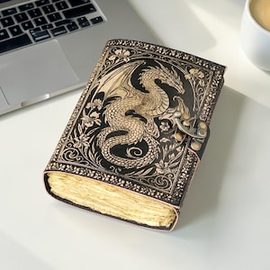 May include: A black leather journal with a detailed dragon design embossed on the cover. The dragon is tan, with intricate wing and scale details. The journal has a metal clasp and aged paper pages. A laptop and a cup of coffee are in the background.