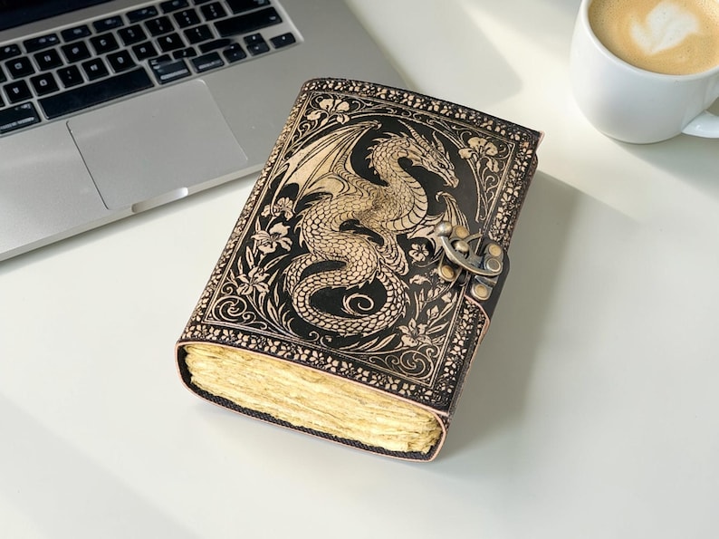 May include: A black leather journal with a detailed dragon design embossed on the cover. The dragon is tan, with intricate wing and scale details. The journal has a metal clasp and aged paper pages. A laptop and a cup of coffee are in the background.
