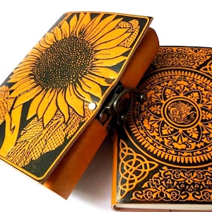 Könnte beinhalten: Zwei Lederjournale mit aufwendigen Designs. Eines zeigt eine Sonnenblume, das andere ein keltisches Knotenmuster. Die Journale sind in Gelb- und Schwarztönen gehalten und haben einen Metallverschluss. Die Journale sind wahrscheinlich handgefertigt.