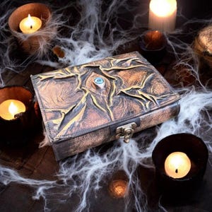 May include: A brown and gold leather-bound book with an embossed eye design and a clasp closure. The book is surrounded by lit candles and cobwebs, creating a mystical and gothic aesthetic. The eye has a blue center.