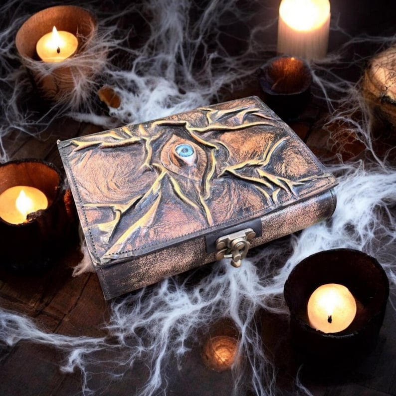 May include: A brown and gold leather-bound book with an embossed eye design and a clasp closure. The book is surrounded by lit candles and cobwebs, creating a mystical and gothic aesthetic. The eye has a blue center.