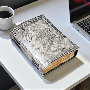 May include: A silver-colored journal with a dragon design on the cover, next to a white coffee mug with black coffee and a laptop. The journal has intricate floral and dragon details. The pages are aged.