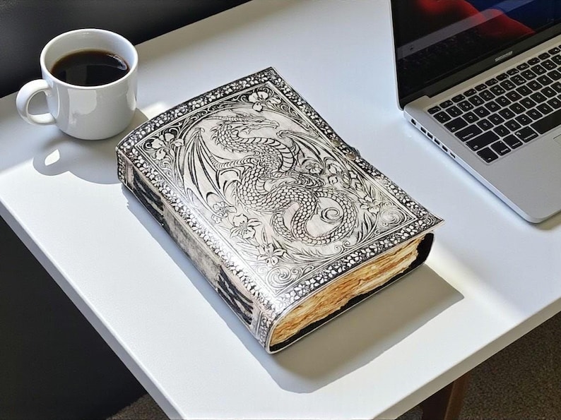 May include: A silver-colored journal with a dragon design on the cover, next to a white coffee mug with black coffee and a laptop. The journal has intricate floral and dragon details. The pages are aged.