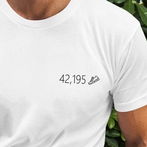 以下が含まれることがあります： 「42,195」の文字と、ランニングシューズの小さなグラフィックがプリントされた白いTシャツです。