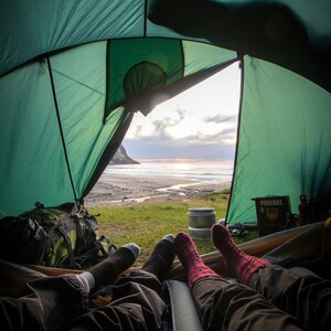 K&ouml;nnte beinhalten: Zwei Personen entspannen in einem gr&uuml;nen Zelt mit Blick auf einen Strand und das Meer. Das Zelt ist offen und der Himmel hat eine wundersch&ouml;ne Sonnenuntergangsfarbe. Die Personen tragen Socken und Hosen.