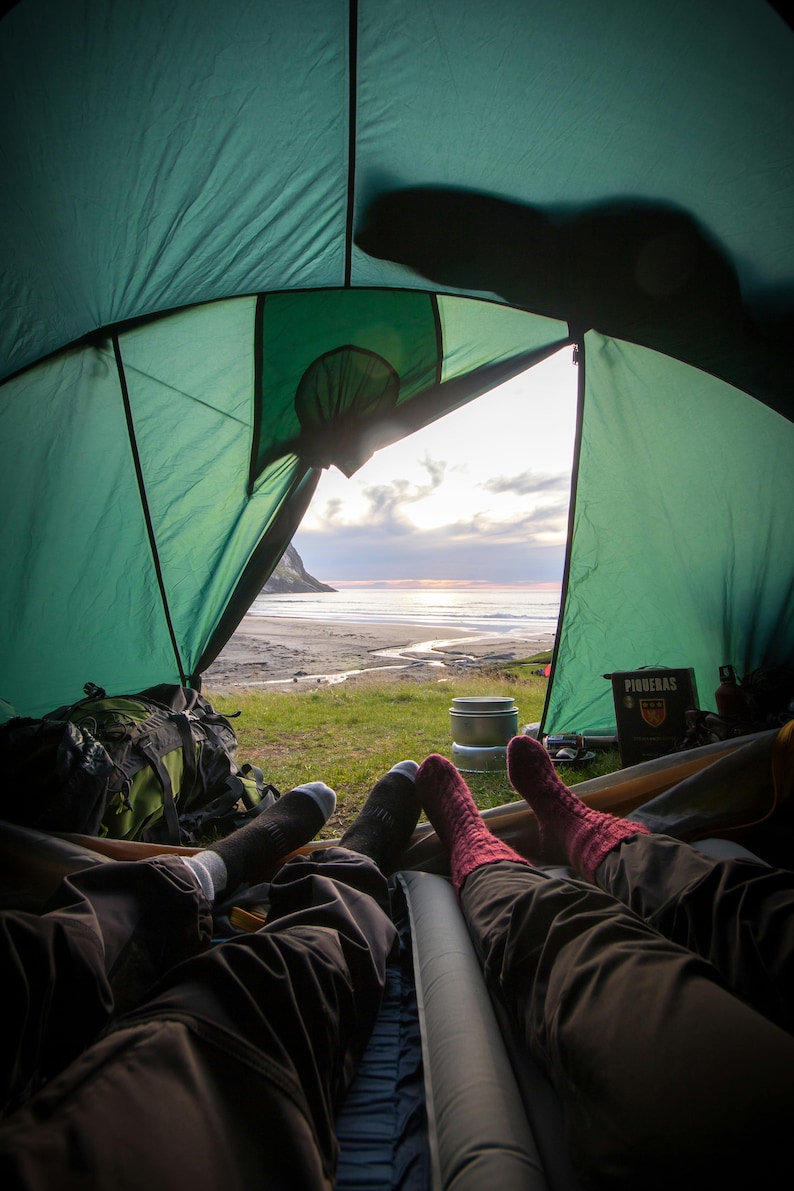 K&ouml;nnte beinhalten: Zwei Personen entspannen in einem gr&uuml;nen Zelt mit Blick auf einen Strand und das Meer. Das Zelt ist offen und der Himmel hat eine wundersch&ouml;ne Sonnenuntergangsfarbe. Die Personen tragen Socken und Hosen.