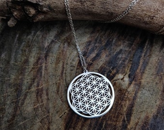 May include: A silver necklace with a flower of life pendant. The pendant is a circular design with intricate details. The necklace is on a wooden surface.