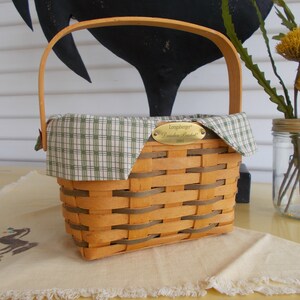 May include: A rectangular woven basket with a wooden handle and a green plaid fabric lining. A small brass plaque reads "Longaberger Dresden Basket 2001". The basket sits on a yellow surface.
