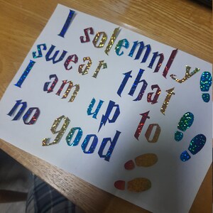 May include: A white card featuring the quote "I solemnly swear I am up to no good" in iridescent, glittery lettering. The design includes glittery footprints and a lightning bolt.