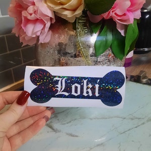 May include: A white bone-shaped sticker with the name "Loki" in a gothic font. The bone is filled with a dark blue glitter design. The background includes a vase of pink and yellow flowers.