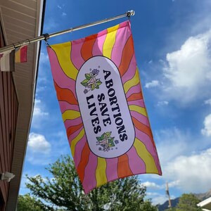 Puede incluir: Una bandera rosa y amarilla con un diseño de rayos de sol y el texto "ABORTIONS SAVE LIVES" en un óvalo blanco.