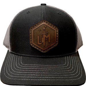 Könnte beinhalten: Eine schwarze und graue Trucker-Kappe mit einem braunen Leder-Patch, auf dem ein Schwert und die Buchstaben "UM" in einer sechseckigen Form zu sehen sind.