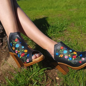 May include: Black peep-toe heeled sandals with a floral embroidery design in shades of blue, red, yellow, and green.