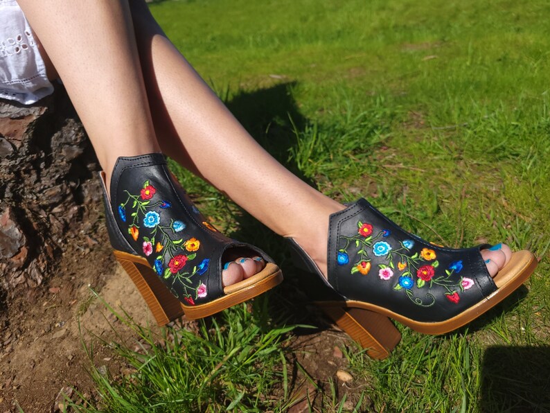 May include: Black peep-toe heeled sandals with a floral embroidery design in shades of blue, red, yellow, and green.