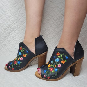 May include: Black peep-toe heeled sandals with a floral embroidery design. The embroidery features colourful flowers in shades of red, orange, blue, and green.
