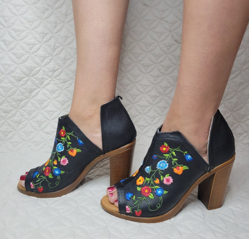 May include: Black peep-toe heeled sandals with a floral embroidery design. The embroidery features colourful flowers in shades of red, orange, blue, and green.