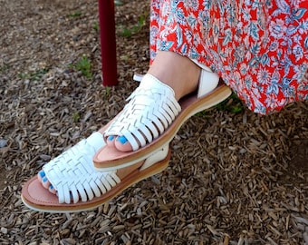 May include: White leather sandals with woven straps and a brown sole. The sandals are worn on a person's feet with blue nail polish.