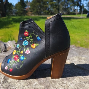 May include: Black leather peep-toe heeled bootie with colourful floral embroidery. The bootie has a wooden heel and a peep-toe design.