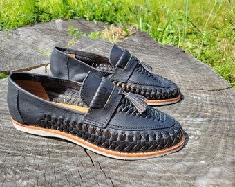 May include: A pair of black leather loafers featuring a woven texture on the vamp and sides, a fringed tongue detail, and a decorative tassel. The shoes have a tan interior lining, a natural-colored leather welt, and a white rubber outsole.
