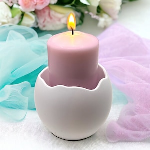 Può includere: Una candela rosa con un portacandele in ceramica bianca a forma di uovo. La candela è accesa e la fiamma è visibile. La candela è su una superficie bianca con tessuto blu e rosa.
