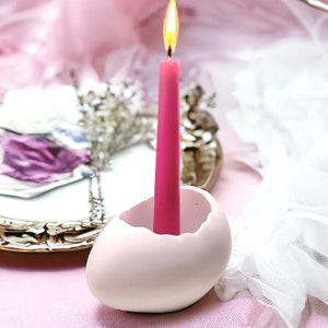Może przedstawiać: Biały ceramiczny świecznik w kształcie jajka z różową świecą w środku. Świeca jest zapalona i płomień jest widoczny.