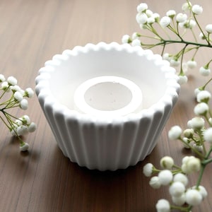 Può includere: Un portacandele in ceramica bianca con un design scanalato e un centro circolare. Il portacandele è circondato da fiori bianchi e poggia su una superficie di legno. Il portacandele è un oggetto decorativo.