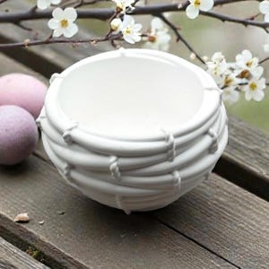 Op de afbeelding: Een witte keramische kom met een textuur, touwachtig ontwerp rond de buitenkant. De kom is rond met een brede opening. Twee bolvormige badbruisballen, één roze en één paars, liggen naast de kom. Witte bloemen en takken zijn op de achtergrond.