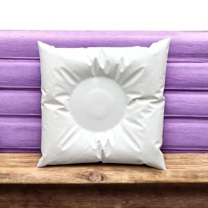 May include: A square white pillow with a circular indentation in the center. The pillow is made of a soft, textured material and is resting on a wooden surface. The background features a purple wooden wall.