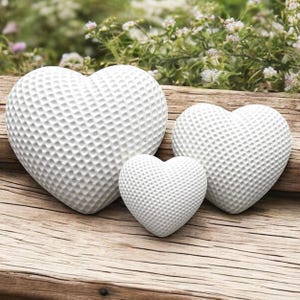 Op de afbeelding: Drie witte hartvormige decoratieve objecten met een textuuroppervlak, gerangschikt op een houten oppervlak. De harten variëren in grootte, met de grootste links en de kleinste in het midden. De achtergrond toont groen.