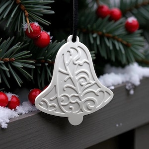 Puede incluir: Adorno navideño blanco en forma de campana con un intrincado diseño floral. Cuelga de un cordón negro sobre un fondo de ramas de hoja perenne y bayas rojas, evocando un tema festivo de vacaciones.