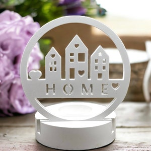 Könnte beinhalten: Weißer Kerzenhalter aus Holz mit einem runden Design, das drei Häuser und das Wort "HOME" mit Herzen zeigt. Der Kerzenhalter hat eine kleine Öffnung für ein Teelicht.