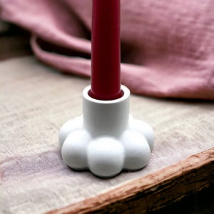 Könnte beinhalten: Weißer Kerzenhalter aus Keramik mit abgerundetem, bauchigem Sockel. Der Halter hat eine einzige Öffnung oben für eine Kerze. Eine rote Kerze befindet sich im Halter.