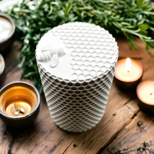 Può includere: Una candela bianca cilindrica con un motivo a nido d'ape e un disegno di ape sulla parte superiore. La candela è circondata da piccole candele accese e verde, il tutto su una superficie di legno rustico. Il design della candela evoca un tema naturale e organico.