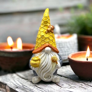 Könnte beinhalten: Eine gelbe und goldene Gnomfigur mit einem Honigwabenhut, die einen Honigtopf mit der Aufschrift "Honey" und einen Honiglöffel hält. Der Gnom hat einen weißen Bart und einen grauen Körper.