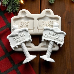 Könnte beinhalten: Eine weiße Silikonform für die Herstellung von Weihnachtsdekorationen. Die Form zeigt zwei Schilder: eines mit der Aufschrift "North Pole" und das andere mit "Merry Christmas". Die Schilder sind auf Pfosten mit Schneedesign montiert.