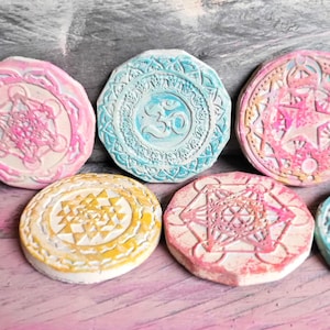 May include: Six round, white, ceramic coasters with colourful, intricate designs. The designs include a mandala, a star, and the symbol Om.