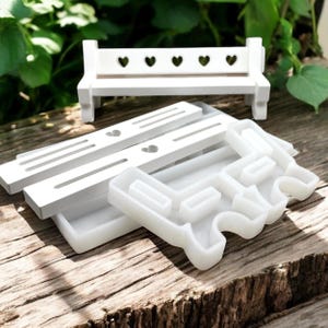 Könnte beinhalten: Eine Sammlung weißer Miniaturbänke und Formen. Die Bank hat eine Lattenlehne mit herzförmigen Aussparungen. Mehrere weiße Formen befinden sich im Vordergrund, mit einem herzförmigen Ausschnitt oben. Auf einer Holzoberfläche.