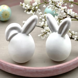 Könnte beinhalten: Zwei weiße Keramik-Hasenfiguren mit langen Ohren, auf einem hellrosa Tablett. Die Hasen sind eiförmig, wobei die Ohren die Oberseite bilden. Dekorative Ostereier und weiße Blumen sind im Hintergrund.