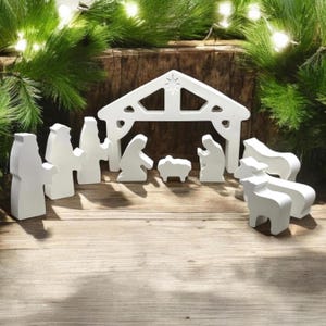 Op de afbeelding: Wit houten kerststal met een stal, Maria, Jozef, baby Jezus, een schaap en drie herders. De figuren staan ​​allemaal op een houten oppervlak.