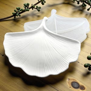 Può includere: Piatti da portata bianchi a forma di foglia. I piatti presentano un motivo a venature dettagliato e un bordo curvo e smerlato. Il design ricorda una foglia di ginkgo. I piatti sono su una superficie di legno.