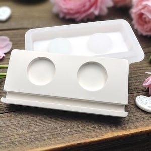 Peut inclure: Un moule en silicone blanc pour le bricolage, avec deux cavités circulaires et une forme rectangulaire. Le moule est ouvert, révélant un couvercle assorti avec deux inserts circulaires. Le design est posé sur une surface en bois.