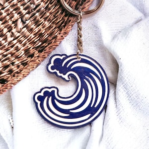 Könnte beinhalten: Ein blauer und weißer Wellen-Schlüsselanhänger mit einer goldenen Kette. Die Welle ist stilisiert und hat einen weißen Umriss.