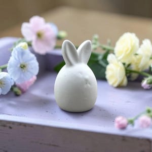 Könnte beinhalten: Kleine, weiße Keramik-Hasenfigur mit aufrechten Ohren. Der Hase ist eiförmig und sitzt auf einem hellvioletten Tablett, umgeben von pastellfarbenen Blumen. Eine bezaubernde Oster- oder Frühlingsdekoration.